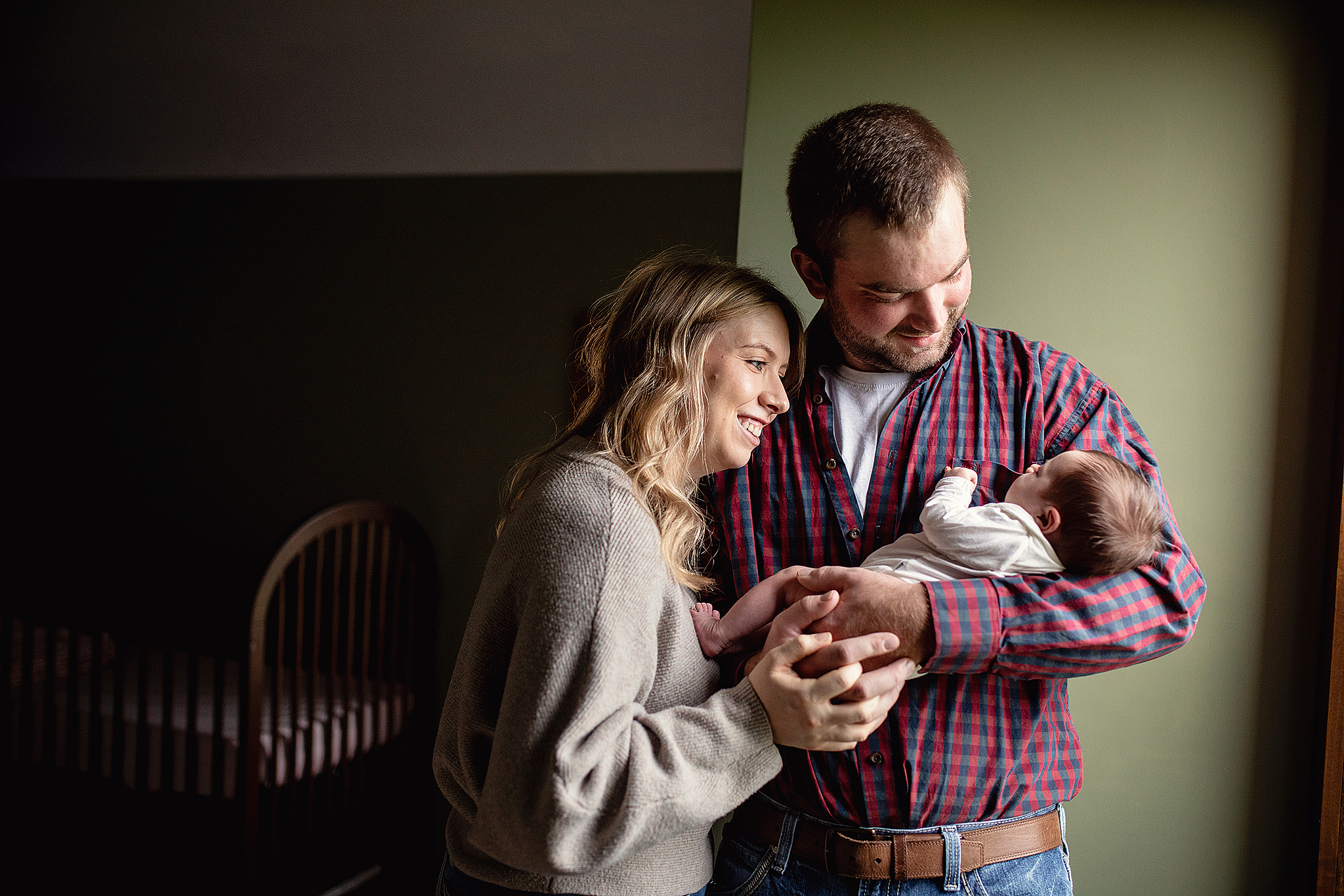 Dad and mom holding newborn baby standing in front of window in babies nursery
