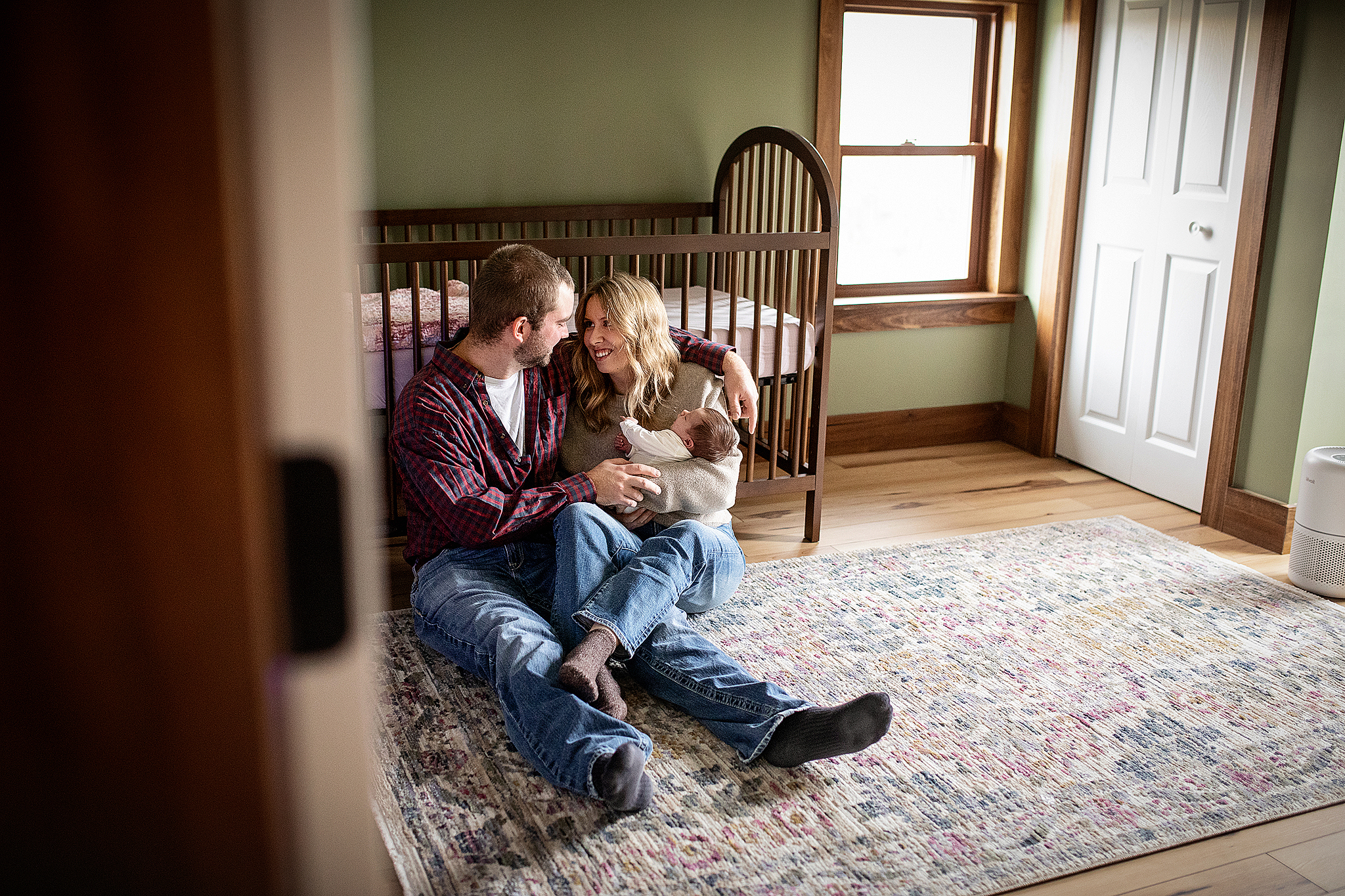 peaking in the nursery of a newborn baby. Mom and dad sitting close to each other cuddling baby. In home newborn session