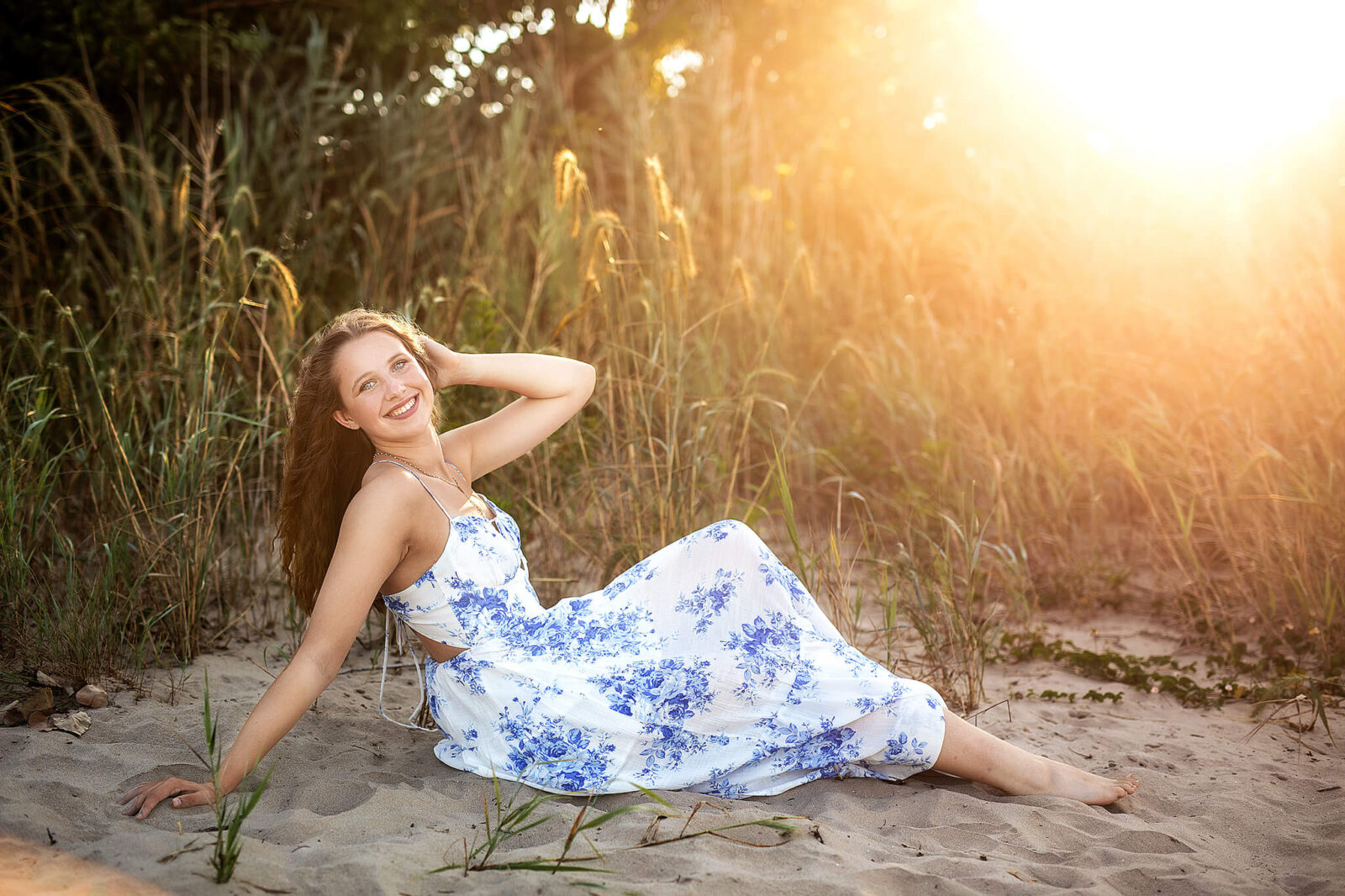 High school senior girl sitting on sandy beach with beach sand and sun behind her