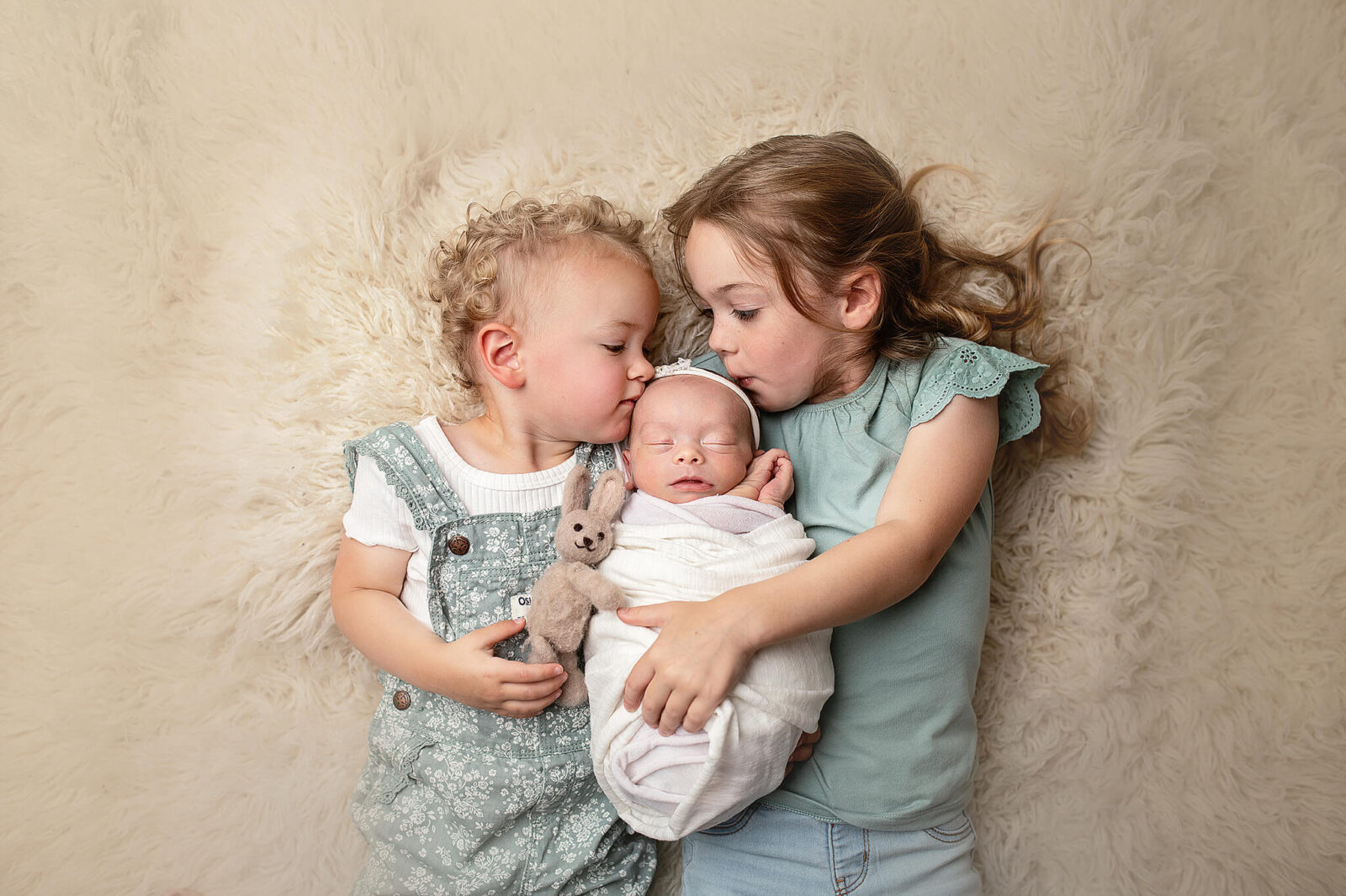 Two older siblings kissing the head of a newborn baby sister in a studio for newborn photography in bucyrus ohio