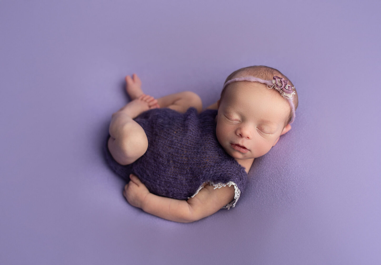 newborn baby girl in a purple outfit curled on a purple backdrop